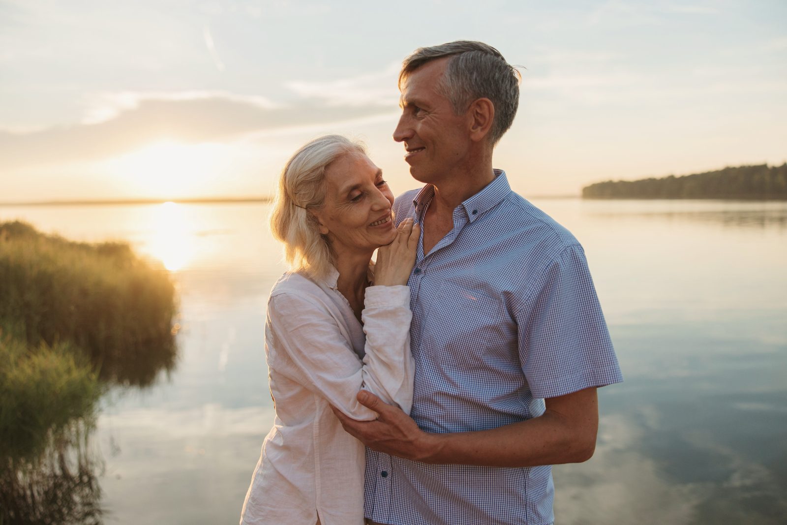 A gray-haired couple holding each other by the water, in golden light